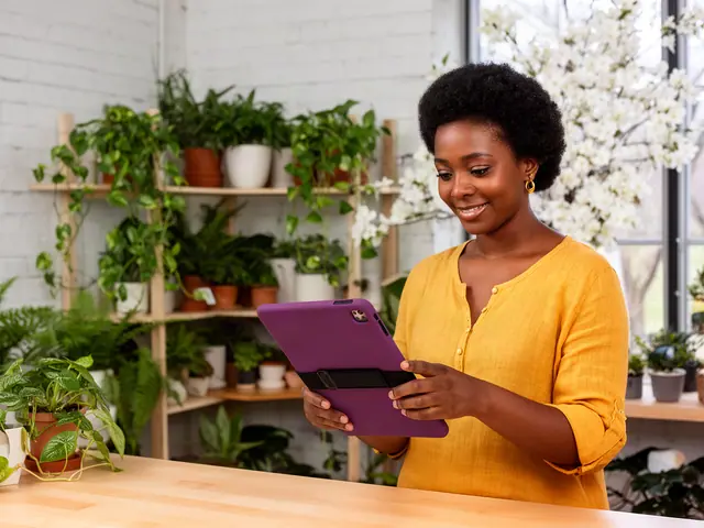 Plant shop owner in a mustard top smiles at a tablet among shelves of potted houseplants